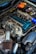 A close-up view of a car engine with various parts visible, including a large air filter, hoses, and a blue valve cover. The image also shows an orange battery and numerous wires and components arranged around the engine bay.
