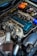 A close-up view of a car engine with various parts visible, including a large air filter, hoses, and a blue valve cover. The image also shows an orange battery and numerous wires and components arranged around the engine bay.