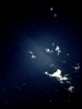A night sky filled with stars, featuring clouds illuminated by a soft light. The darkness is pierced by tiny, sparkling stars scattered across the scene, with clouds adding texture and depth.