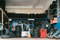 A garage filled with tires and automotive equipment. Tires are stacked on shelves and some are mounted on the walls alongside wheel rims. A person is seated in the corner on the left, looking at a mobile phone. An orange traffic cone is on the floor, and there is a machine used for tire services.