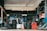 A garage filled with tires and automotive equipment. Tires are stacked on shelves and some are mounted on the walls alongside wheel rims. A person is seated in the corner on the left, looking at a mobile phone. An orange traffic cone is on the floor, and there is a machine used for tire services.