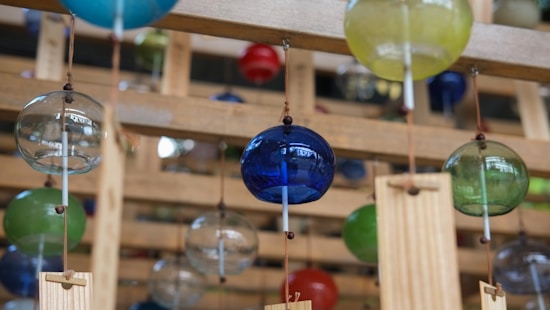 A collection of colorful glass wind chimes hangs from wooden structures. The glass spheres come in shades of blue, green, yellow, and clear, attached by strings with metal rods. The wooden backdrop creates a warm, rustic ambiance.