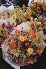 A collection of vibrant floral bouquets wrapped in paper, featuring a rich variety of flowers and foliage including purple, pink, and yellow blooms alongside textured leaves.
