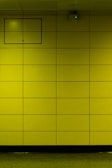 A yellow tiled wall with a small rectangular access panel and a security camera mounted near the ceiling. The tiles are arranged in a grid, and there is a reflective, polished floor at the bottom.