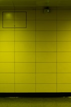 A yellow tiled wall with a small rectangular access panel and a security camera mounted near the ceiling. The tiles are arranged in a grid, and there is a reflective, polished floor at the bottom.