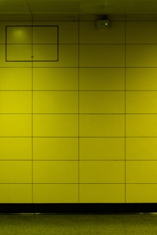 A yellow tiled wall with a small rectangular access panel and a security camera mounted near the ceiling. The tiles are arranged in a grid, and there is a reflective, polished floor at the bottom.