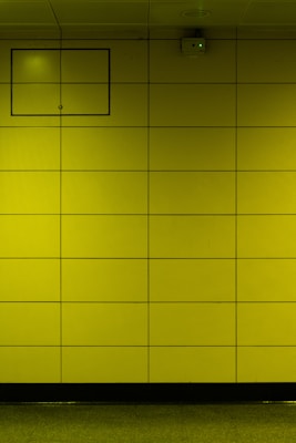 A yellow tiled wall with a small rectangular access panel and a security camera mounted near the ceiling. The tiles are arranged in a grid, and there is a reflective, polished floor at the bottom.