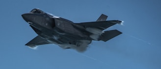 A military jet is flying at high speed through the sky. Vapor trails can be seen near its wings, indicating rapid movement. The aircraft has a sleek and modern design.