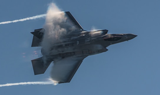 An advanced fighter jet is captured mid-flight against a clear blue sky, with vapor trailing from its wings indicating high speed. The jet's powerful engines emit a faint orange glow.
