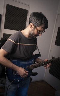 A person is playing an electric guitar in a room with soundproofing panels on the walls. The individual is wearing glasses and a casual t-shirt with colorful stripes. There is a focus on the concentration of the person while playing the instrument.