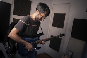 A person with short, dark hair and glasses is playing an electric guitar. The individual is standing in a room with soundproofing panels on the walls, suggesting a music or recording studio environment. The guitar is a deep blue color, and the person is focused on playing, with their head tilted downward.