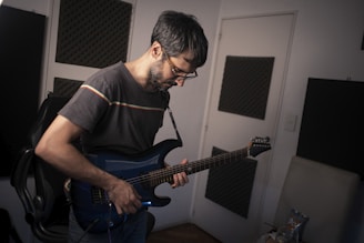 A person with short, dark hair and glasses is playing an electric guitar. The individual is standing in a room with soundproofing panels on the walls, suggesting a music or recording studio environment. The guitar is a deep blue color, and the person is focused on playing, with their head tilted downward.