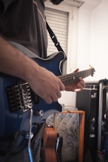 A person is playing a blue electric guitar in a room. The guitar has a sleek design with a tremolo bridge. There are other string instruments and equipment visible in the background, such as a guitar case and an amplifier. The setting appears to be casual, likely a practice or rehearsal space with a window in the background.
