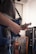 A person is playing a blue electric guitar in a room. The guitar has a sleek design with a tremolo bridge. There are other string instruments and equipment visible in the background, such as a guitar case and an amplifier. The setting appears to be casual, likely a practice or rehearsal space with a window in the background.
