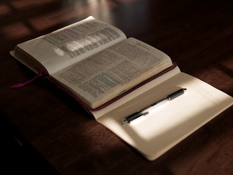 An open Bible with highlighted passages lies on a wooden table, partially illuminated by warm sunlight. Beside it, a blank notebook with lined pages holds a black pen, casting a subtle shadow. The scene conveys a sense of calm and contemplation.