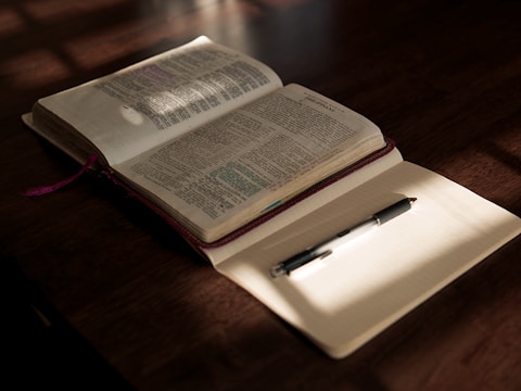 An open Bible with highlighted passages lies on a wooden table, partially illuminated by warm sunlight. Beside it, a blank notebook with lined pages holds a black pen, casting a subtle shadow. The scene conveys a sense of calm and contemplation.