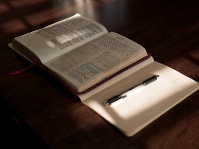 An open Bible with highlighted passages lies on a wooden table, partially illuminated by warm sunlight. Beside it, a blank notebook with lined pages holds a black pen, casting a subtle shadow. The scene conveys a sense of calm and contemplation.
