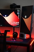 A dual-monitor setup on a desk with one monitor in landscape orientation and the other in portrait. A camera is mounted above one monitor. The desktop wallpaper features a bold, abstract design with red and black colors. The desk is illuminated with red LED lights, and there are various items like a mouse, keyboard, and other gadgets.