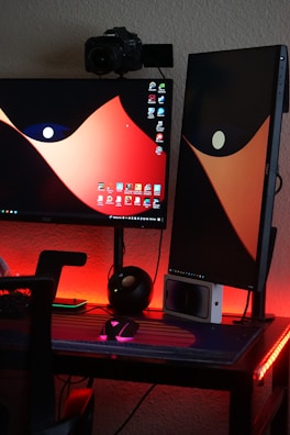 A dual-monitor setup on a desk with one monitor in landscape orientation and the other in portrait. A camera is mounted above one monitor. The desktop wallpaper features a bold, abstract design with red and black colors. The desk is illuminated with red LED lights, and there are various items like a mouse, keyboard, and other gadgets.
