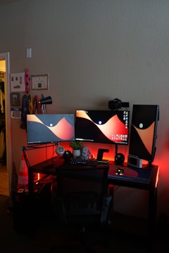 A computer setup featuring three monitors displaying an abstract wallpaper with red and black hues. The desk is equipped with a keyboard, a small potted plant, a microphone, and an elephant figurine. Red LED lights illuminate the area, adding an atmospheric glow. A gaming chair is positioned in front of the desk, and a camera is mounted on one of the monitor stands.