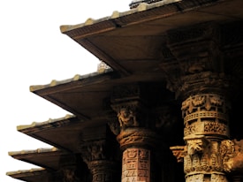 Ornate stone columns feature intricate carvings and decorative patterns, part of an ancient architectural structure. The stonework showcases fine craftsmanship with detailed sculptures and motifs engraved into the columns.