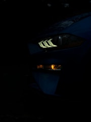 A close-up of a car's headlight in a low-light setting, showing distinct angular LED lights and a lower indicator light glowing amber. The dark background emphasizes the illuminated features of the vehicle.