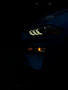 A close-up of a car's headlight in a low-light setting, showing distinct angular LED lights and a lower indicator light glowing amber. The dark background emphasizes the illuminated features of the vehicle.