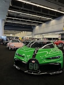 A vibrant green sports car with sleek aerodynamic design is prominently displayed indoors, amidst a line of other luxury vehicles. The space is an exhibition area with high ceilings and industrial lighting. The floor is covered in dark carpet, adding contrast to the vibrant colors of the cars. Several people are in the background, suggesting a public automotive event.