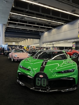A vibrant green sports car with sleek aerodynamic design is prominently displayed indoors, amidst a line of other luxury vehicles. The space is an exhibition area with high ceilings and industrial lighting. The floor is covered in dark carpet, adding contrast to the vibrant colors of the cars. Several people are in the background, suggesting a public automotive event.