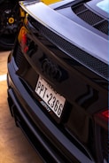 A close-up view of the rear of a black Audi car featuring a Texas license plate with the number PPT 2366. The design includes a sleek, sporty finish with prominent Audi logo and rear grille. In the background, a yellow sports car is partially visible, identifiable by its wheel rim and logo.