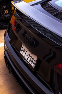 A close-up view of the rear of a black Audi car featuring a Texas license plate with the number PPT 2366. The design includes a sleek, sporty finish with prominent Audi logo and rear grille. In the background, a yellow sports car is partially visible, identifiable by its wheel rim and logo.
