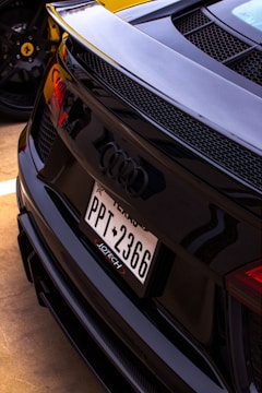 A close-up view of the rear of a black Audi car featuring a Texas license plate with the number PPT 2366. The design includes a sleek, sporty finish with prominent Audi logo and rear grille. In the background, a yellow sports car is partially visible, identifiable by its wheel rim and logo.
