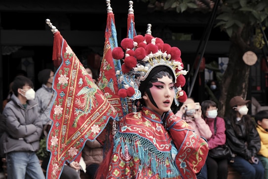 A person adorned in traditional Chinese opera attire is performing outdoors. The elaborate costume features vibrant red fabric with intricate designs, including embroidered floral and phoenix motifs. The headdress is adorned with large, red pompoms and ornate details. Several people in the background are observing the performance, some wearing masks and holding cameras.