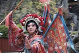 A performer dressed in traditional Chinese opera attire featuring vibrant red, blue, and gold colors. The outfit is adorned with intricate embroidery and patterns, and the headdress is elaborate with floral and ornate designs. The performer holds a decorative stick and appears to be in mid-performance against a backdrop of blurred foliage, suggesting an outdoor setting.