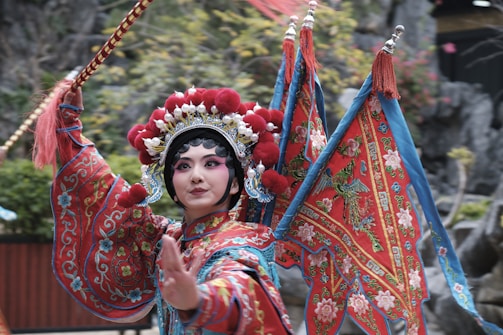 A performer dressed in traditional Chinese opera attire featuring vibrant red, blue, and gold colors. The outfit is adorned with intricate embroidery and patterns, and the headdress is elaborate with floral and ornate designs. The performer holds a decorative stick and appears to be in mid-performance against a backdrop of blurred foliage, suggesting an outdoor setting.