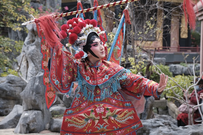 A performer is wearing traditional Chinese opera attire, which is richly adorned with intricate embroidery and vibrant colors. The individual is posing with one hand extended and the other holding a stick decorated with red tassels. The background features a garden setting with rocks and foliage.