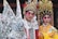 Two people dressed in traditional Chinese opera costumes stand side by side. The ornate costumes feature intricate patterns, vibrant colors, and elaborate headpieces adorned with jewels and feathers. Both individuals have bold, theatrical makeup emphasizing their eyes and eyebrows.
