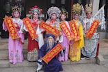 A group of people are dressed in elaborate, colorful traditional costumes. They hold bright red banners with intricate gold designs and Chinese characters. Their attire includes detailed headpieces, vibrant robes, and makeup that enhances the traditional aesthetic.