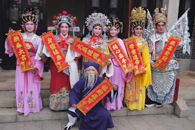 A group of people are dressed in elaborate, colorful traditional costumes. They hold bright red banners with intricate gold designs and Chinese characters. Their attire includes detailed headpieces, vibrant robes, and makeup that enhances the traditional aesthetic.
