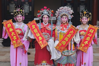 Four individuals dressed in traditional, colorful opera costumes are holding red and gold banners with Chinese characters. The costumes are ornate, featuring intricate embroidery and headdresses adorned with flowers and beads. They stand in front of a traditional building with dark wooden doors, exuding a sense of cultural celebration.