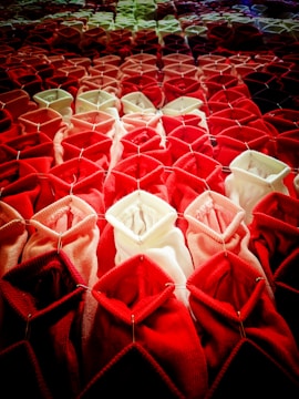 Rows of fabric or textile structures arranged in a gradient from dark red at the bottom to lighter pink and white towards the top. Each piece of fabric is folded in a geometric pattern, creating a three-dimensional, tactile appearance. The fabric appears to be secured with metal pins, adding a structural element.