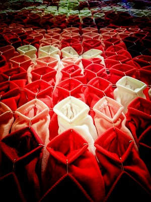 Rows of fabric or textile structures arranged in a gradient from dark red at the bottom to lighter pink and white towards the top. Each piece of fabric is folded in a geometric pattern, creating a three-dimensional, tactile appearance. The fabric appears to be secured with metal pins, adding a structural element.