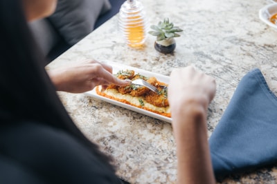 A person using a fork is about to eat a dish consisting of breaded food items placed over a base of what seems to be sauce, with some garnish on top. The background features a textured stone or marble table, a small potted succulent plant, and a honey dispenser.