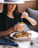 A woman in a wide-brimmed hat is seated at a table enjoying a meal. She holds a cocktail garnished with fruit and appears to be squeezing a fruit peel into her drink. The plate in front of her contains waffles topped with fried chicken, cucumber slices, and sauce. The setting is warm and inviting, with a mix of natural and artificial light creating a cozy atmosphere.
