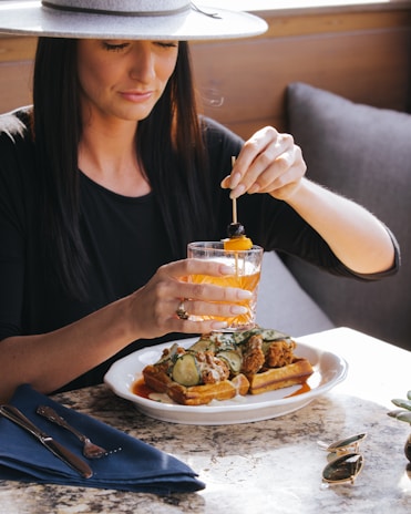A woman in a wide-brimmed hat is seated at a table enjoying a meal. She holds a cocktail garnished with fruit and appears to be squeezing a fruit peel into her drink. The plate in front of her contains waffles topped with fried chicken, cucumber slices, and sauce. The setting is warm and inviting, with a mix of natural and artificial light creating a cozy atmosphere.