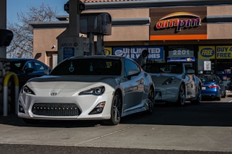 Two sports cars parked at a gas station, one of which is a white Scion FR-S. They are positioned near an ARCO fuel pump with an AMPM convenience store in the background. The store displays various promotional signs, and the area is well-shaded with natural light illuminating the scene.