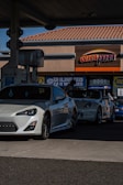 Two sporty white cars are parked at a gas station with the ampm sign visible in the background. The station appears to be a typical service area with facilities for fuel and possible car washes.