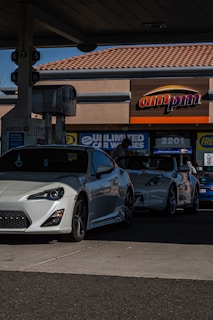 Two sporty white cars are parked at a gas station with the ampm sign visible in the background. The station appears to be a typical service area with facilities for fuel and possible car washes.