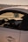 A close-up view of a car’s interior from the passenger side, the dashboard and steering wheel are visible. A decorative item hangs from the rearview mirror. The exterior is sleek and shiny, and in the background, a hazy sky and distant treetops can be seen.