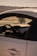 A close-up view of a car’s interior from the passenger side, the dashboard and steering wheel are visible. A decorative item hangs from the rearview mirror. The exterior is sleek and shiny, and in the background, a hazy sky and distant treetops can be seen.